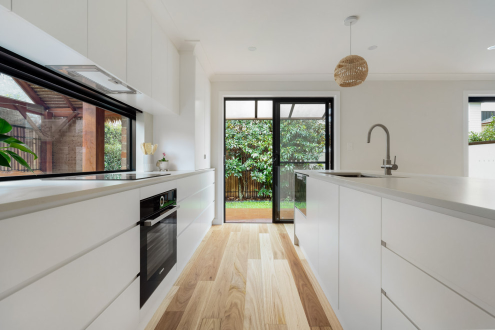 This is an example of a mid-sized beach style galley open plan kitchen in Sydney with an undermount sink, flat-panel cabinets, white cabinets, quartz benchtops, window splashback, black appliances, medium hardwood floors, with island and white benchtop.