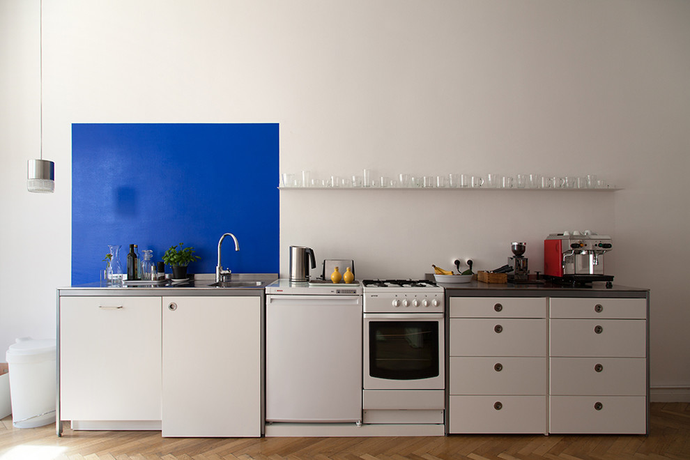 This is an example of a small contemporary single-wall open plan kitchen in Berlin with an integrated sink, flat-panel cabinets, white cabinets, stainless steel benchtops, blue splashback, white appliances, light hardwood floors, no island, limestone splashback and brown floor.