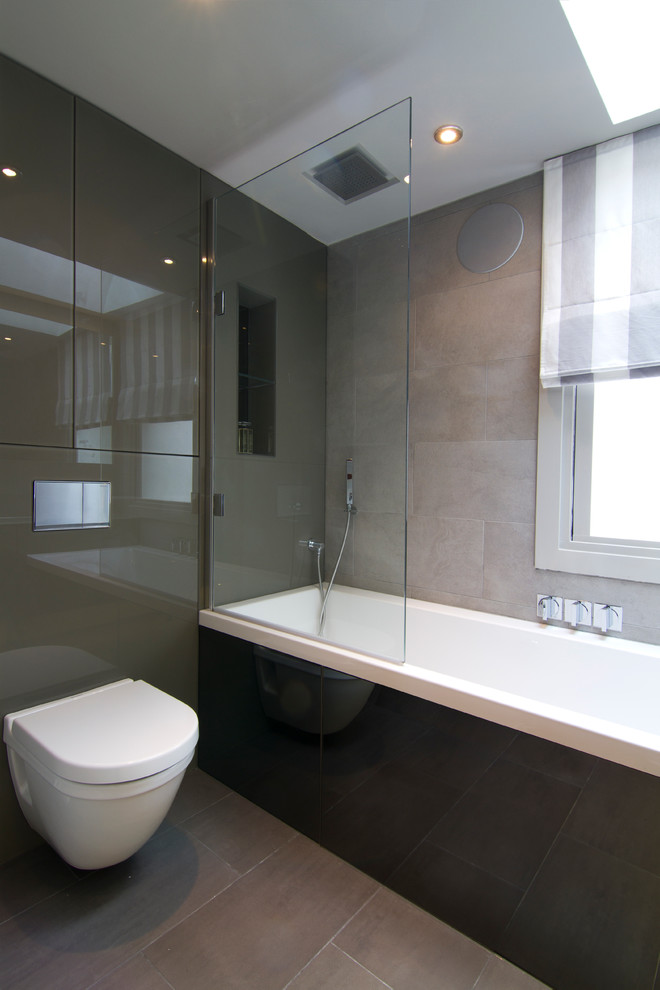 Photo of a small contemporary bathroom in London with a drop-in tub, a shower/bathtub combo, a wall-mount toilet, gray tile, porcelain tile, grey walls and porcelain floors.