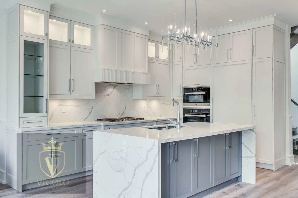 Large transitional l-shaped light wood floor and brown floor kitchen photo with a double-bowl sink, shaker cabinets, white cabinets, quartz countertops, white backsplash, quartz backsplash, paneled appliances, an island and gray countertops