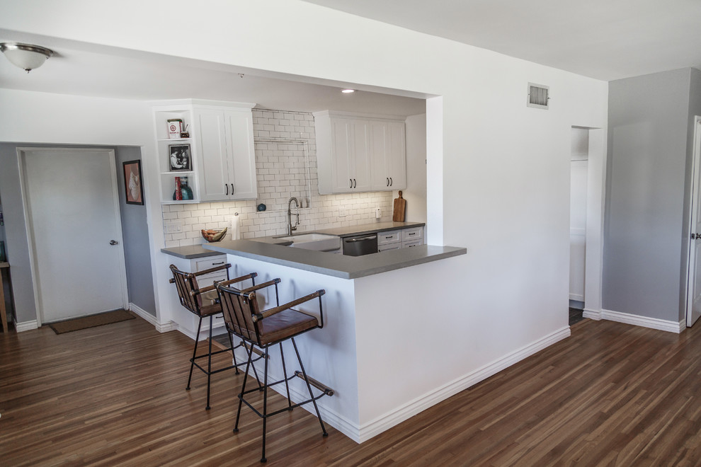 This is an example of a mid-sized contemporary u-shaped separate kitchen in Orange County with a farmhouse sink, shaker cabinets, white cabinets, concrete benchtops, white splashback, subway tile splashback, stainless steel appliances, laminate floors, a peninsula, black floor and grey benchtop.