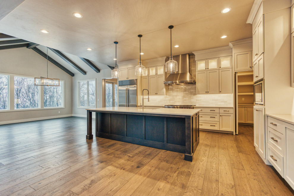 Example of a large transitional l-shaped medium tone wood floor and brown floor kitchen pantry design with an undermount sink, shaker cabinets, white cabinets, quartz countertops, white backsplash, ceramic backsplash, stainless steel appliances, an island and white countertops