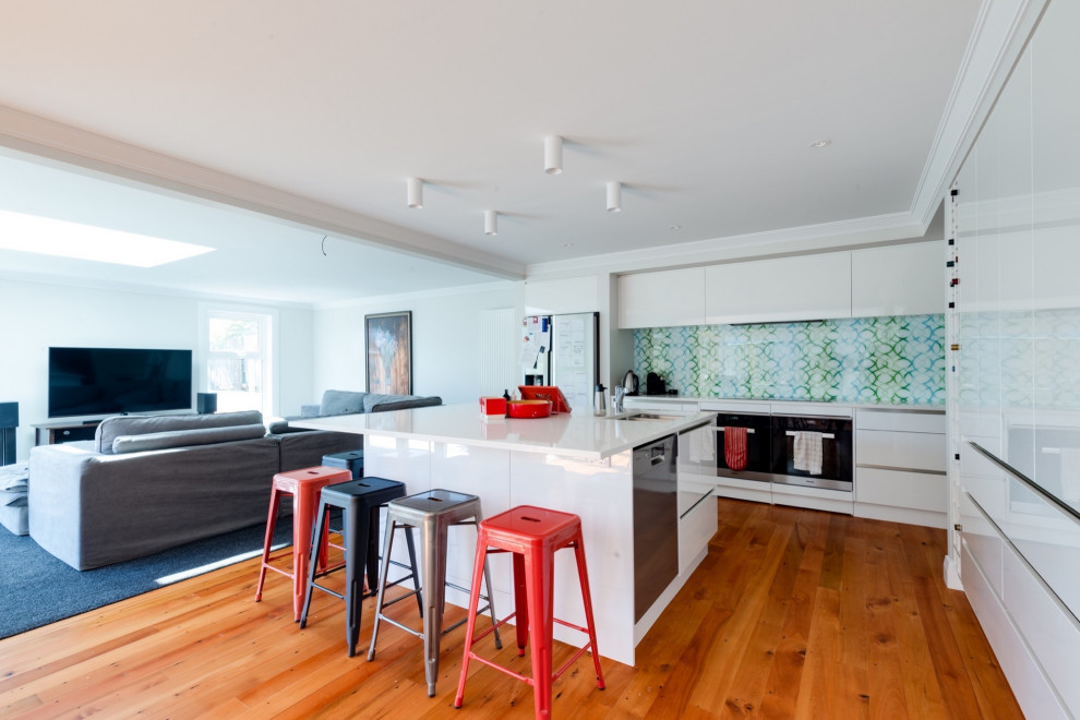 Photo of a large scandinavian galley eat-in kitchen in Sydney with recessed-panel cabinets, white cabinets, granite benchtops, multi-coloured splashback, glass sheet splashback, stainless steel appliances, medium hardwood floors, multiple islands, brown floor and white benchtop.