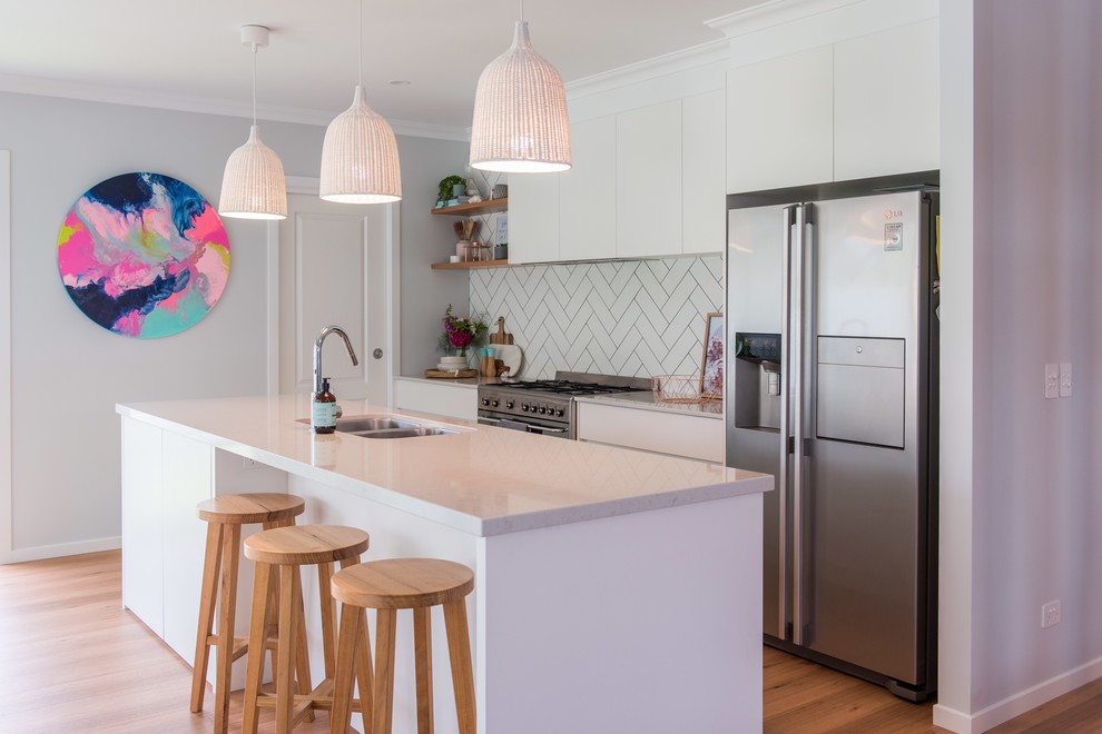 This is an example of a beach style galley eat-in kitchen in Other with an undermount sink, flat-panel cabinets, white cabinets, white splashback, ceramic splashback, stainless steel appliances, medium hardwood floors, with island, brown floor and grey benchtop.