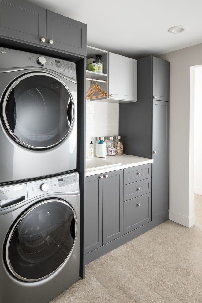 Photo of a transitional single-wall dedicated laundry room in Nashville with a drop-in sink, shaker cabinets, grey cabinets, white walls, a stacked washer and dryer, grey floor and white benchtop.
