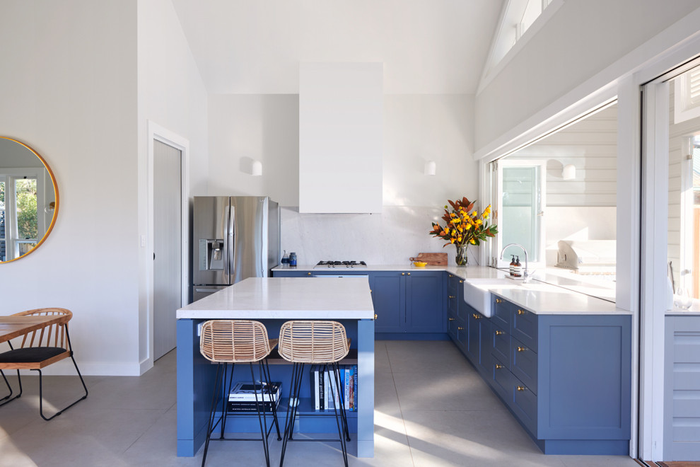 Design ideas for a transitional l-shaped kitchen in Sydney with a farmhouse sink, shaker cabinets, blue cabinets, white splashback, stone slab splashback, with island, grey floor and white benchtop.