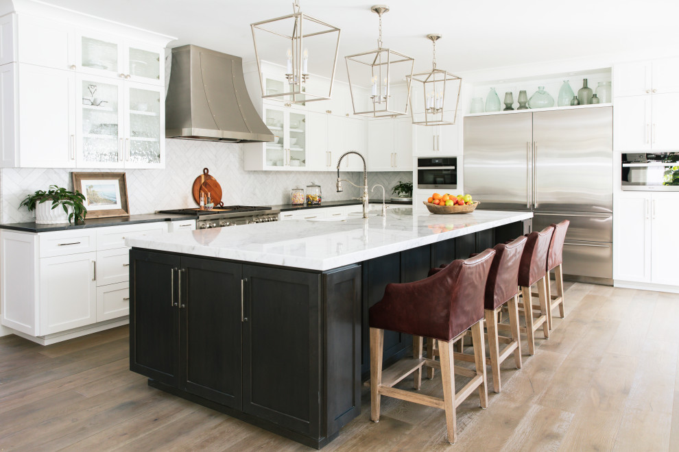 Design ideas for a beach style l-shaped open plan kitchen in San Diego with white cabinets, white splashback, with island, a farmhouse sink, shaker cabinets, stainless steel appliances, medium hardwood floors, brown floor and black benchtop.