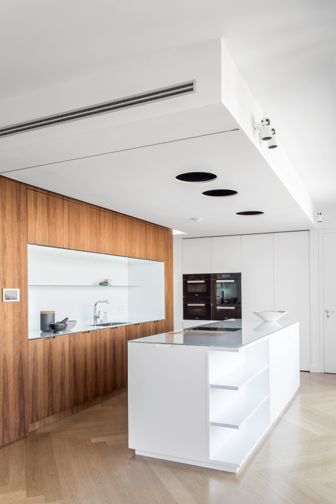 Modern single-wall kitchen in Berlin with a drop-in sink, flat-panel cabinets, white cabinets, glass benchtops, white splashback, black appliances, light hardwood floors and with island.