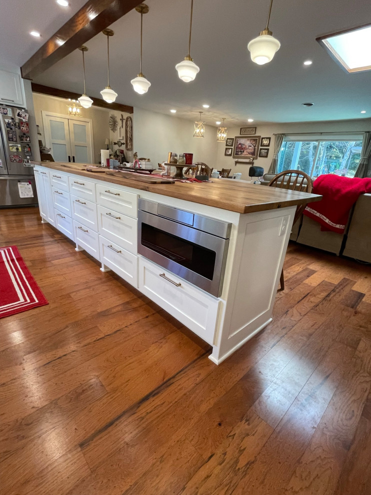 This is an example of a large country l-shaped open plan kitchen in Other with a farmhouse sink, shaker cabinets, white cabinets, quartz benchtops, white splashback, subway tile splashback, white appliances, medium hardwood floors, with island, brown floor and white benchtop.