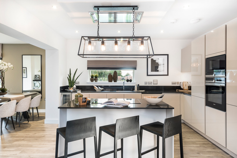 Inspiration for a mid-sized contemporary eat-in kitchen in Hampshire with flat-panel cabinets, beige cabinets, black appliances, light hardwood floors, with island and black splashback.