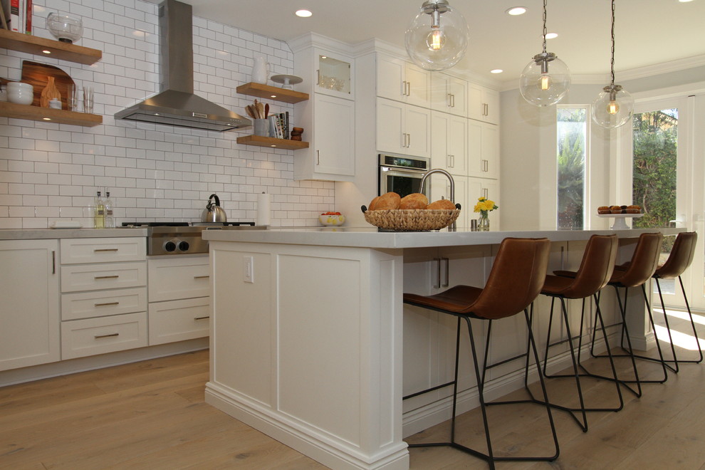 Design ideas for a large transitional l-shaped open plan kitchen in Orange County with a farmhouse sink, shaker cabinets, white cabinets, quartz benchtops, white splashback, subway tile splashback, stainless steel appliances, light hardwood floors, with island and beige floor.