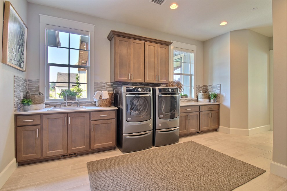 Photo of an expansive arts and crafts l-shaped utility room in Portland with a drop-in sink, flat-panel cabinets, brown cabinets, quartz benchtops, grey walls, ceramic floors, a side-by-side washer and dryer, beige floor and white benchtop.