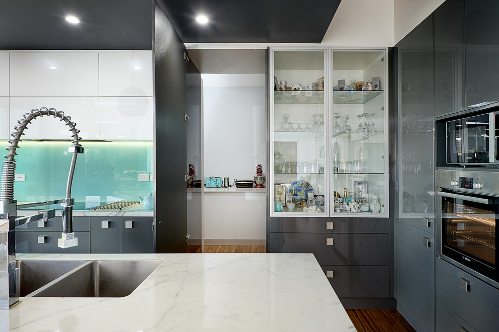 Large contemporary l-shaped eat-in kitchen in Melbourne with a double-bowl sink, flat-panel cabinets, marble benchtops, green splashback, glass sheet splashback, stainless steel appliances, medium hardwood floors and with island.