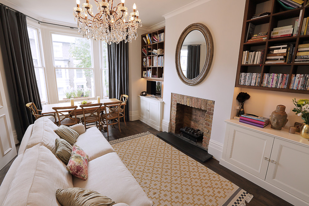 This is an example of a traditional formal open concept living room in London with dark hardwood floors, a standard fireplace, a brick fireplace surround, a freestanding tv and grey walls.