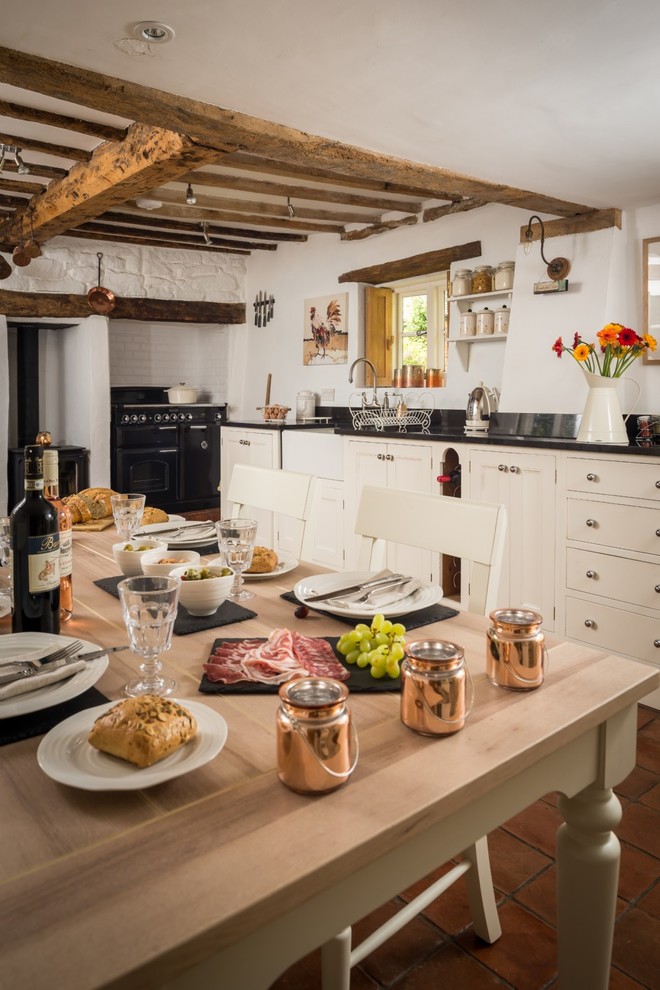 Design ideas for a mid-sized country single-wall eat-in kitchen in Wiltshire with a farmhouse sink, shaker cabinets, white cabinets, laminate benchtops, black splashback, ceramic splashback, black appliances, terra-cotta floors and brown floor.