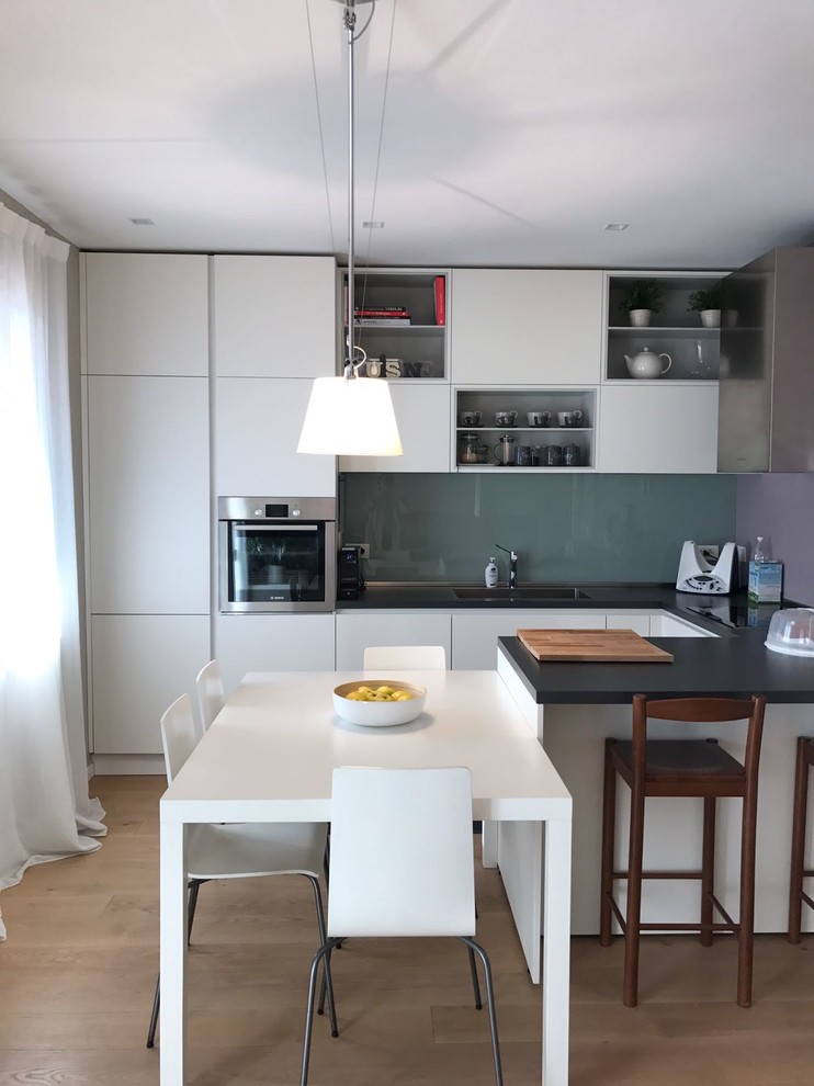Design ideas for a small contemporary single-wall eat-in kitchen in Milan with an integrated sink, flat-panel cabinets, stainless steel cabinets, stainless steel benchtops, grey splashback, glass sheet splashback, stainless steel appliances, light hardwood floors, a peninsula and brown floor.