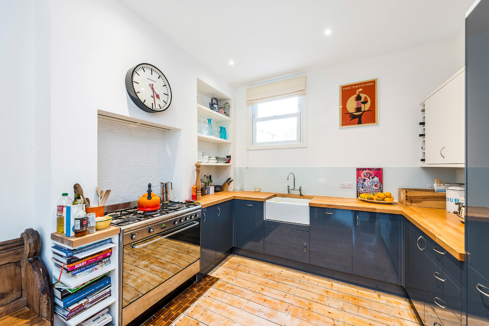 This is an example of a small contemporary u-shaped open plan kitchen in London with a farmhouse sink, flat-panel cabinets, blue cabinets, wood benchtops, white splashback, glass sheet splashback, stainless steel appliances, light hardwood floors, beige floor and brown benchtop.