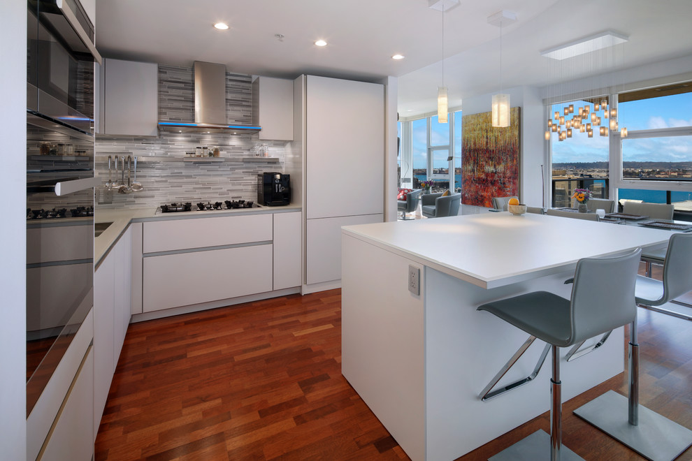 Photo of a contemporary l-shaped kitchen in San Diego with flat-panel cabinets, white cabinets, grey splashback, matchstick tile splashback, panelled appliances, medium hardwood floors, with island, brown floor and white benchtop.
