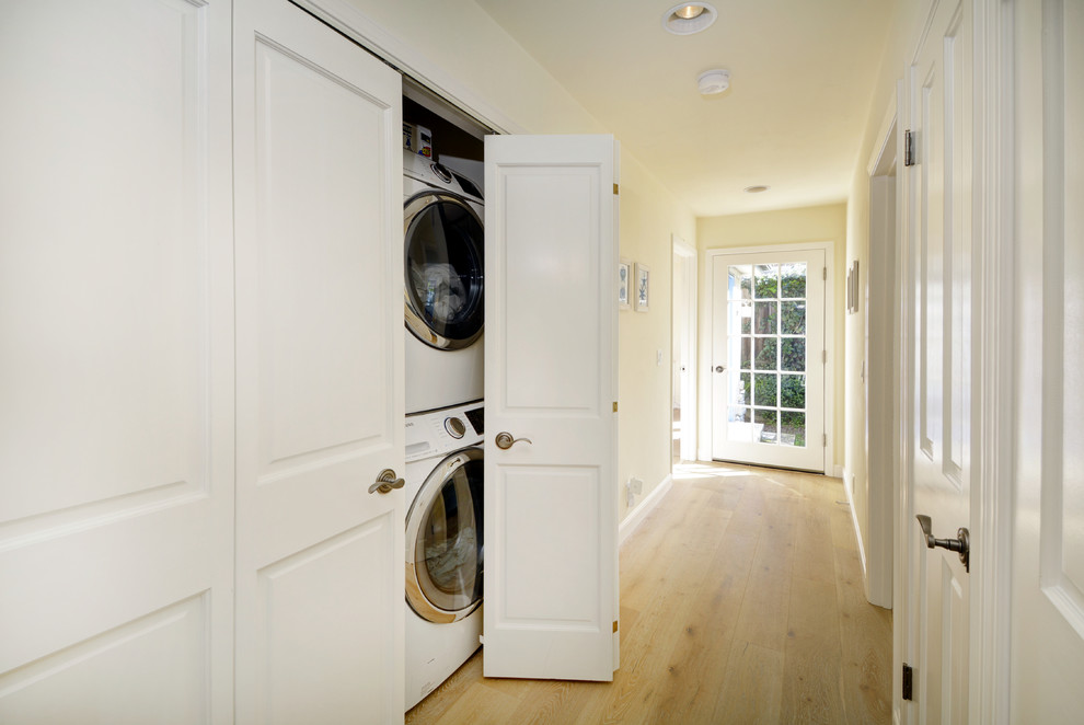 Inspiration for a small country galley laundry cupboard in San Francisco with light hardwood floors, a stacked washer and dryer, white cabinets, yellow walls, brown floor and raised-panel cabinets.