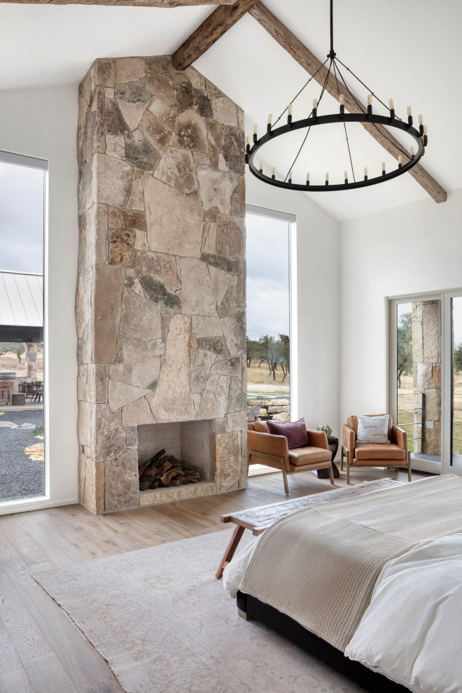 Country master bedroom in Austin with white walls, light hardwood flooring, a standard fireplace, a stone fireplace surround and exposed beams.