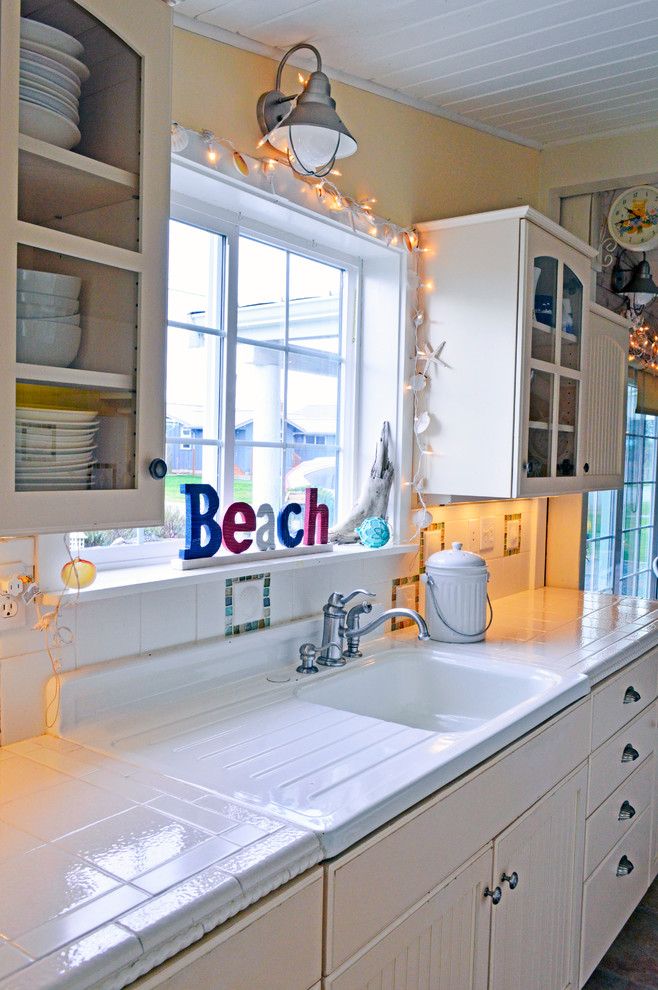 Design ideas for a small beach style galley separate kitchen in Seattle with an integrated sink, recessed-panel cabinets, white cabinets, tile benchtops, white splashback, white appliances, vinyl floors, no island and glass tile splashback.