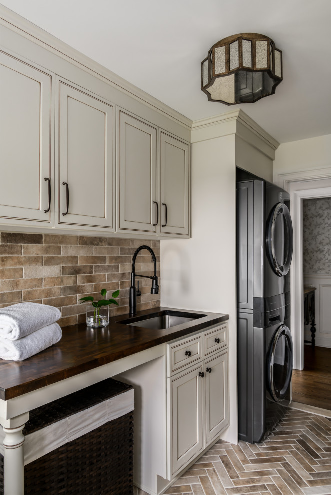 Mid-sized elegant galley ceramic tile and multicolored floor utility room photo in Philadelphia with an undermount sink, shaker cabinets, beige cabinets, wood countertops, subway tile backsplash, beige walls, a stacked washer/dryer and brown countertops
