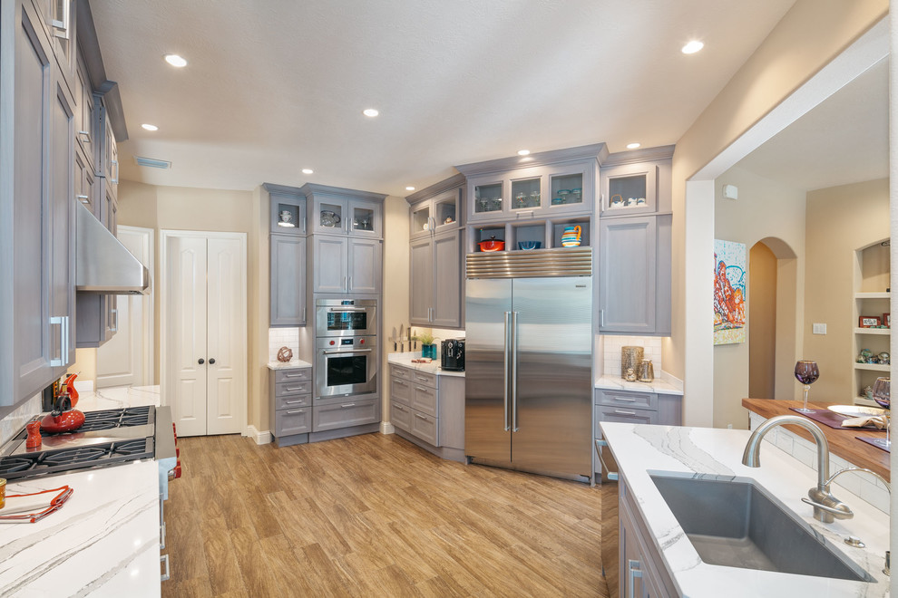 Expansive transitional open plan kitchen in Houston with a single-bowl sink, recessed-panel cabinets, grey cabinets, quartz benchtops, white splashback, subway tile splashback, stainless steel appliances, porcelain floors, with island, brown floor and white benchtop.