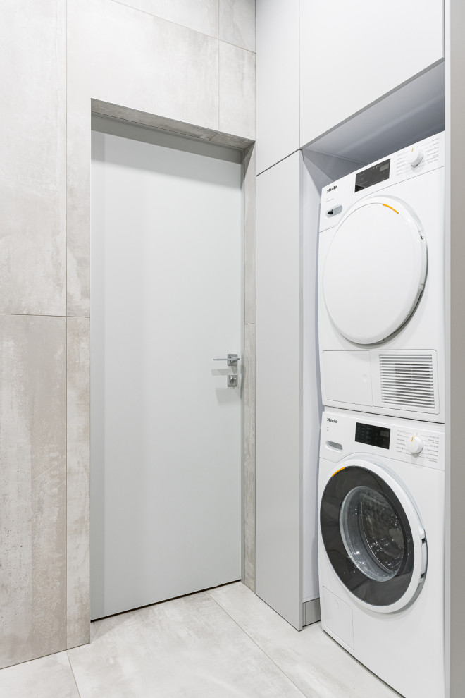 Modelo de lavadero actual con armarios con paneles lisos, puertas de armario grises, paredes grises, suelo de baldosas de porcelana, lavadora y secadora apiladas y suelo gris