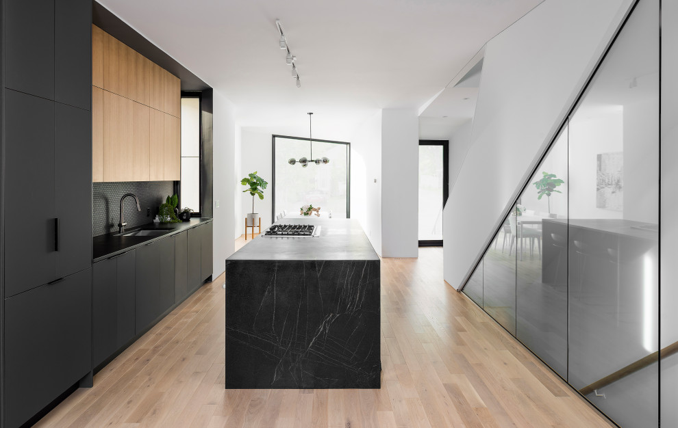 Photo of a modern galley kitchen in Toronto with an undermount sink, flat-panel cabinets, black cabinets, black splashback, mosaic tile splashback, stainless steel appliances, light hardwood floors, with island, beige floor and black benchtop.