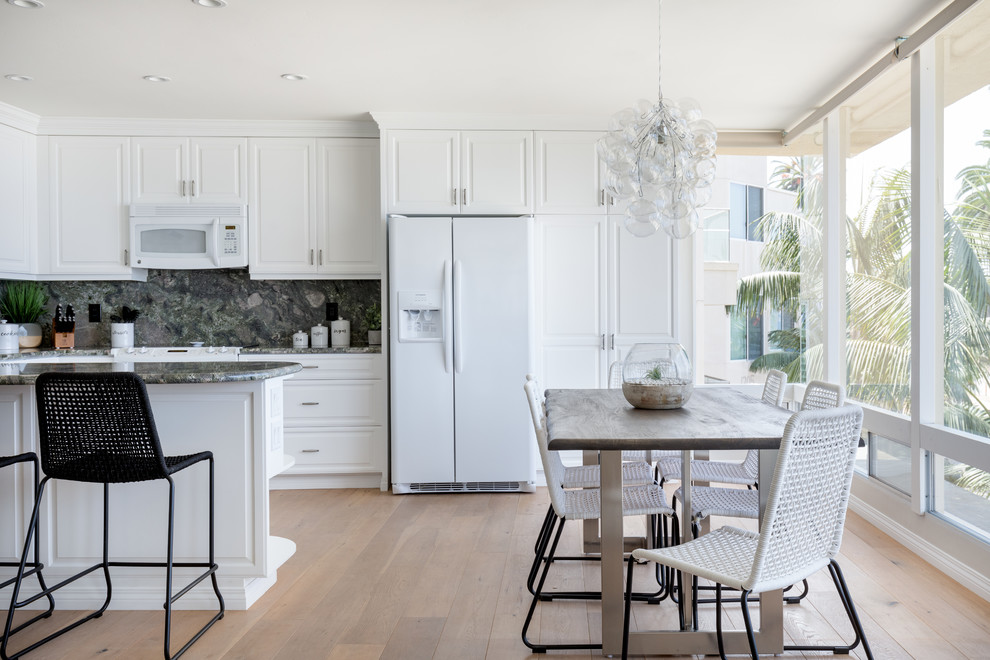Design ideas for a beach style u-shaped eat-in kitchen in San Diego with raised-panel cabinets, white cabinets, grey splashback, white appliances, light hardwood floors, a peninsula, beige floor and grey benchtop.