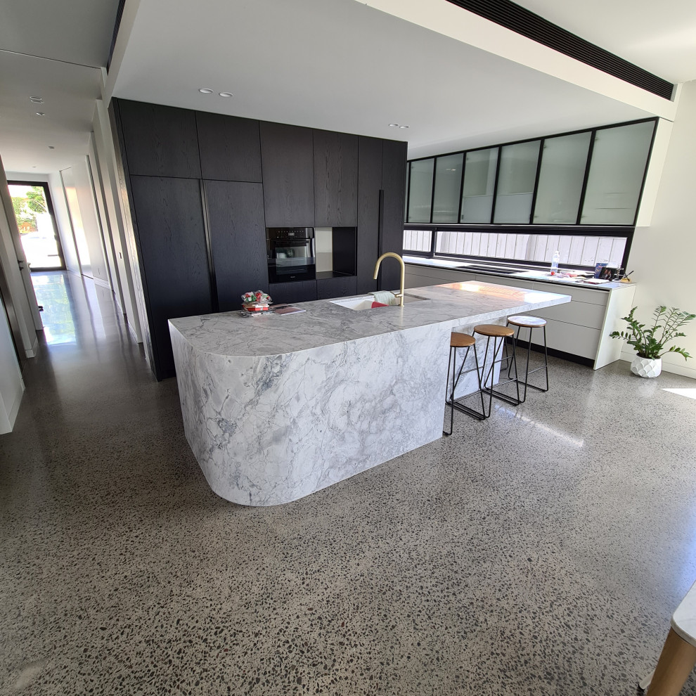 Large contemporary u-shaped open plan kitchen in Melbourne with an undermount sink, flat-panel cabinets, black cabinets, marble benchtops, window splashback, stainless steel appliances, concrete floors, with island, grey floor and white benchtop.