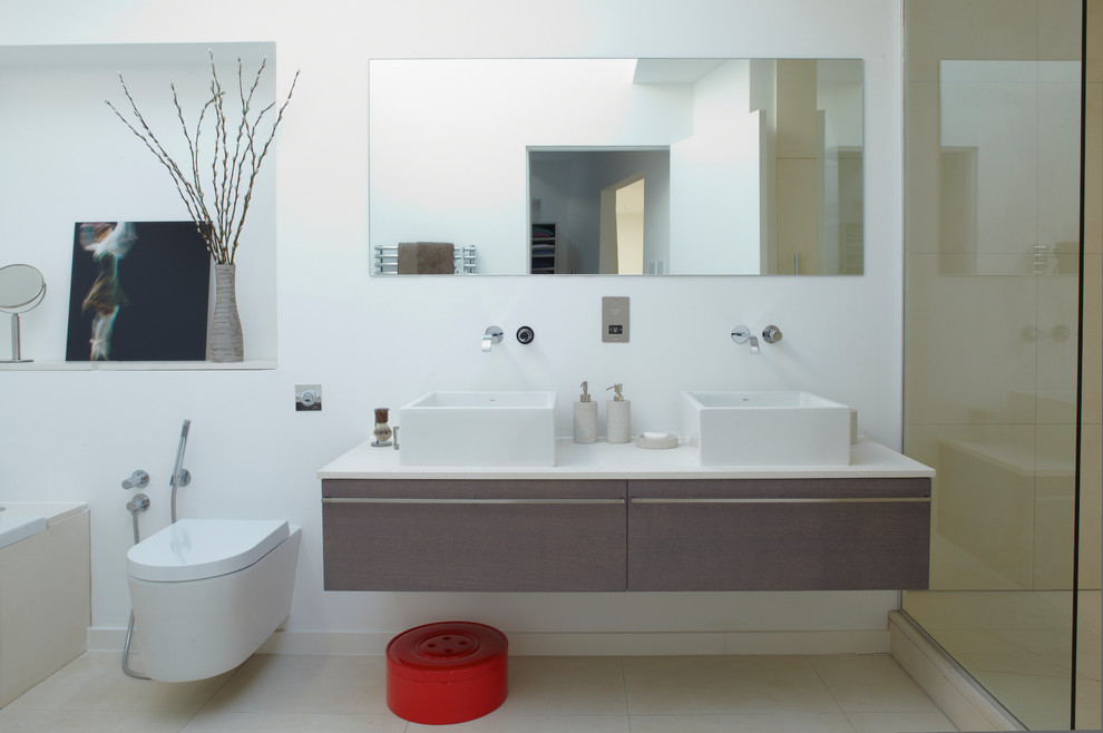 Photo of a mid-sized contemporary master bathroom in London with a vessel sink, flat-panel cabinets, medium wood cabinets, a drop-in tub, a wall-mount toilet, beige tile, white walls and white benchtops.