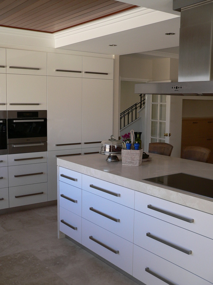 This is an example of a large beach style galley eat-in kitchen in Perth with an undermount sink, white cabinets, marble benchtops, black appliances, travertine floors and with island.
