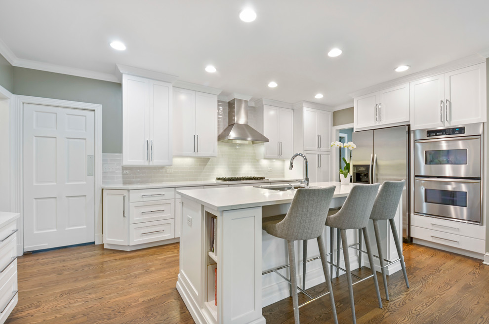 Photo of a mid-sized transitional single-wall open plan kitchen in Chicago with an undermount sink, shaker cabinets, white cabinets, quartz benchtops, grey splashback, ceramic splashback, stainless steel appliances, medium hardwood floors, with island and white benchtop.