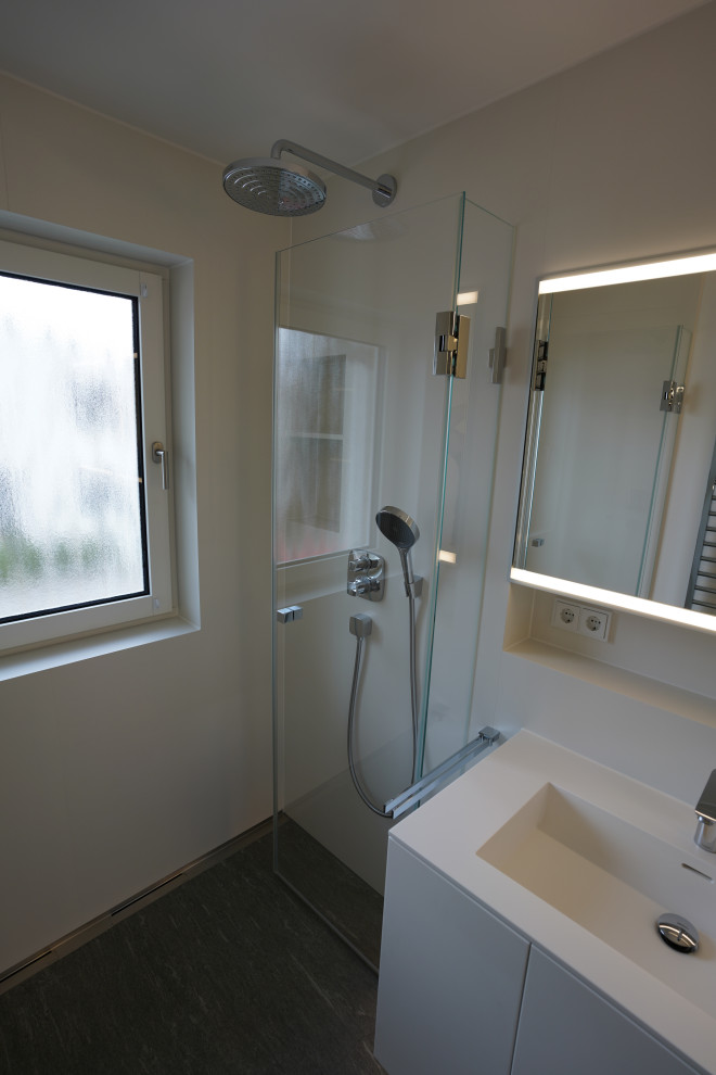 Duschbad With flächenbündigen Schrankfronten, weißen Schränken, bodengleicher Dusche, Wandtoilette, weißen Fliesen, Steinfliesen, Wandwaschbecken, Mineralwerkstoff-Waschtisch, grauem Boden, Falttür-Duschabtrennung, weißer Waschtischplatte, Einzelwaschbecken and schwebendem Waschtisch in Munich
