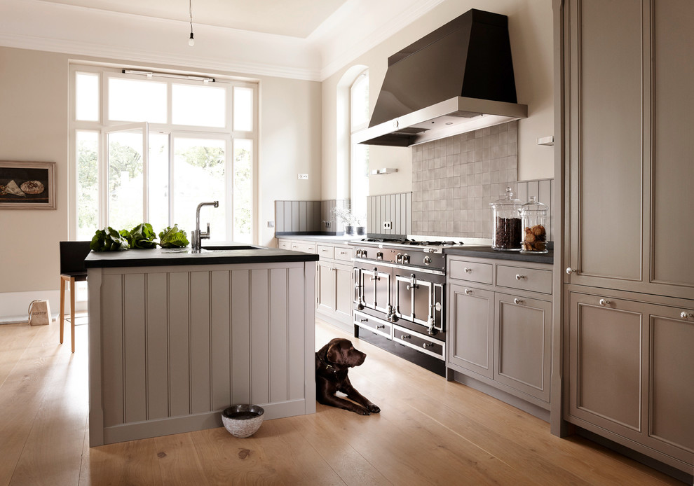 Design ideas for a large country open plan kitchen in Hamburg with an undermount sink, recessed-panel cabinets, grey cabinets, grey splashback, black appliances, light hardwood floors, with island and brown floor.