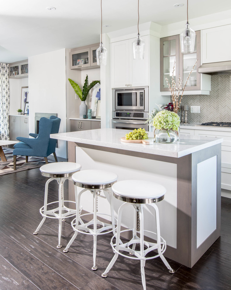 Mid-sized transitional l-shaped open plan kitchen in Toronto with a farmhouse sink, shaker cabinets, white cabinets, quartzite benchtops, grey splashback, stone tile splashback, stainless steel appliances, dark hardwood floors and with island.