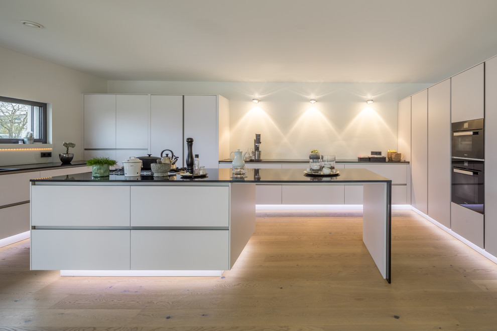 Photo of an expansive contemporary u-shaped open plan kitchen in Other with flat-panel cabinets, white cabinets, granite benchtops, with island, light hardwood floors, brown floor, a drop-in sink, white splashback and black appliances.