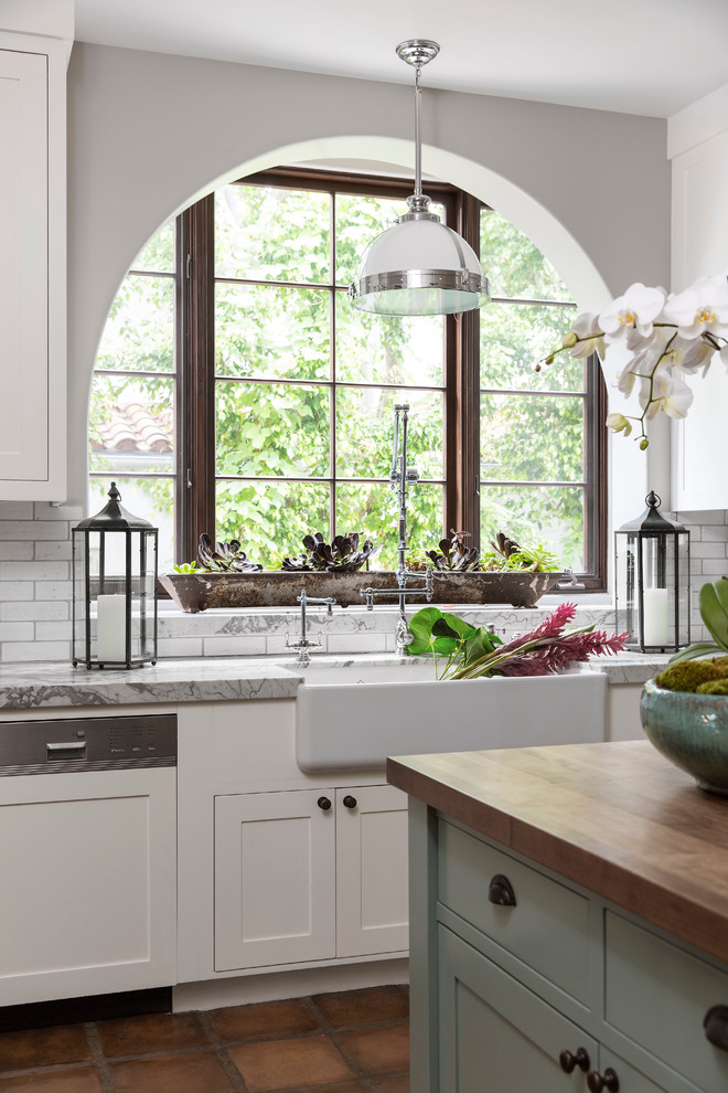 Design ideas for a mediterranean kitchen in Los Angeles with a farmhouse sink, flat-panel cabinets, white cabinets, white splashback, subway tile splashback, stainless steel appliances, terra-cotta floors and with island.