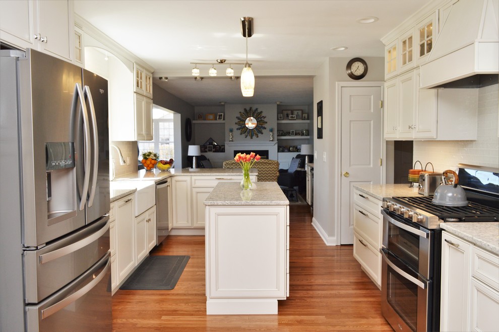 This is an example of a large traditional l-shaped open plan kitchen in Chicago with a farmhouse sink, recessed-panel cabinets, white cabinets, quartzite benchtops, white splashback, glass tile splashback, stainless steel appliances, medium hardwood floors, with island, brown floor and grey benchtop.