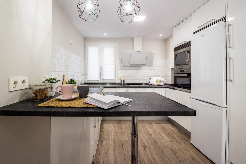 Small contemporary u-shaped separate kitchen in Other with a drop-in sink, flat-panel cabinets, white cabinets, laminate benchtops, laminate floors, with island, black benchtop, white splashback, subway tile splashback, white appliances and brown floor.