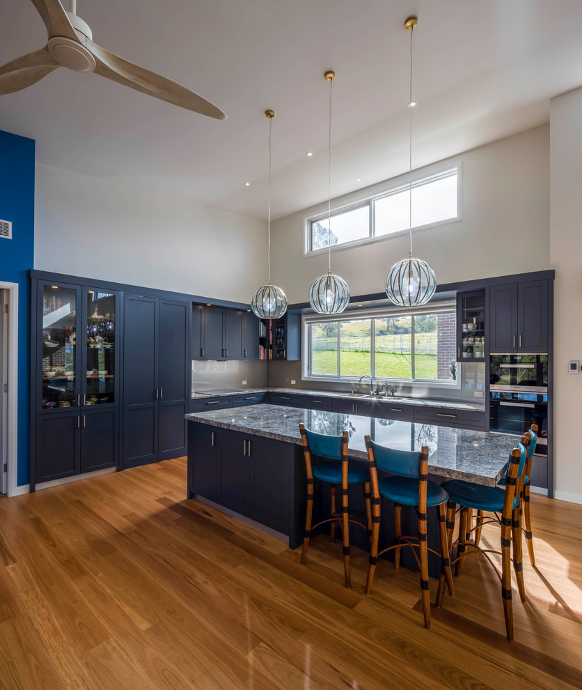 Photo of an expansive contemporary kitchen in Sydney with a double-bowl sink, shaker cabinets, grey cabinets, granite benchtops, metallic splashback, glass sheet splashback, black appliances, light hardwood floors, with island, brown floor and grey benchtop.