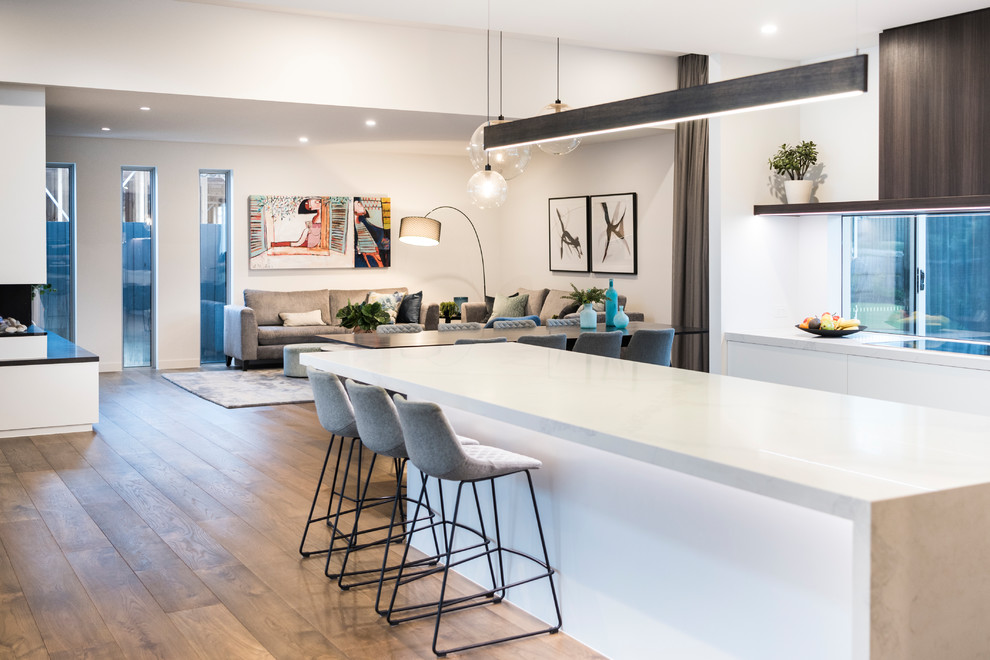This is an example of an expansive contemporary l-shaped open plan kitchen in Melbourne with an undermount sink, quartz benchtops, black appliances, medium hardwood floors, a peninsula, brown floor, white benchtop, flat-panel cabinets, dark wood cabinets and window splashback.