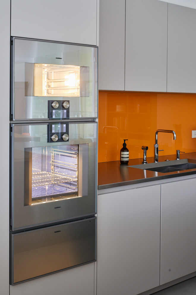 This is an example of a large modern single-wall open plan kitchen with black cabinets, stainless steel benchtops, yellow splashback, glass sheet splashback, stainless steel appliances, ceramic floors, with island and grey floor.