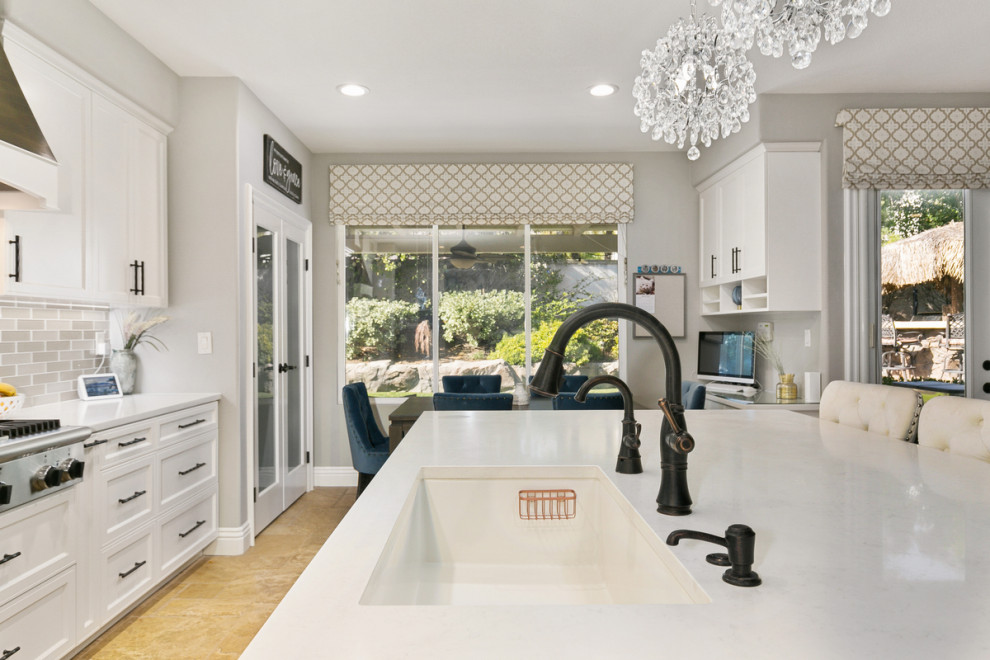 This is an example of an expansive traditional u-shaped open plan kitchen in Orange County with a single-bowl sink, shaker cabinets, white cabinets, engineered stone countertops, multi-coloured splashback, stone tiled splashback, stainless steel appliances, travertine flooring, an island, beige floors and white worktops.