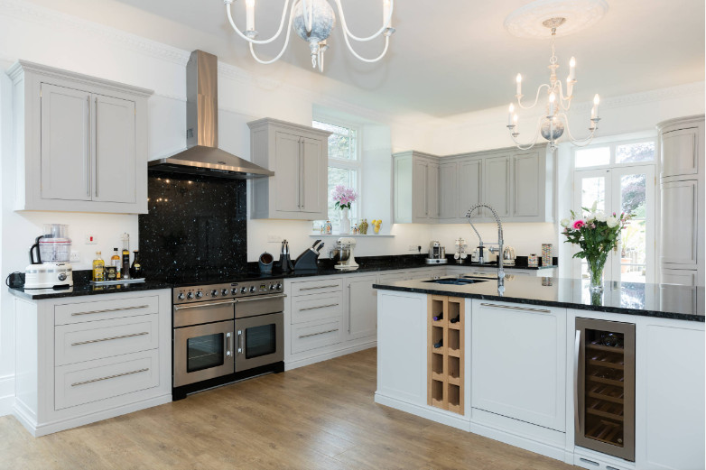Photo of an expansive traditional l-shaped open plan kitchen in Cornwall with an undermount sink, recessed-panel cabinets, grey cabinets, black splashback, panelled appliances, medium hardwood floors, with island, brown floor and black benchtop.