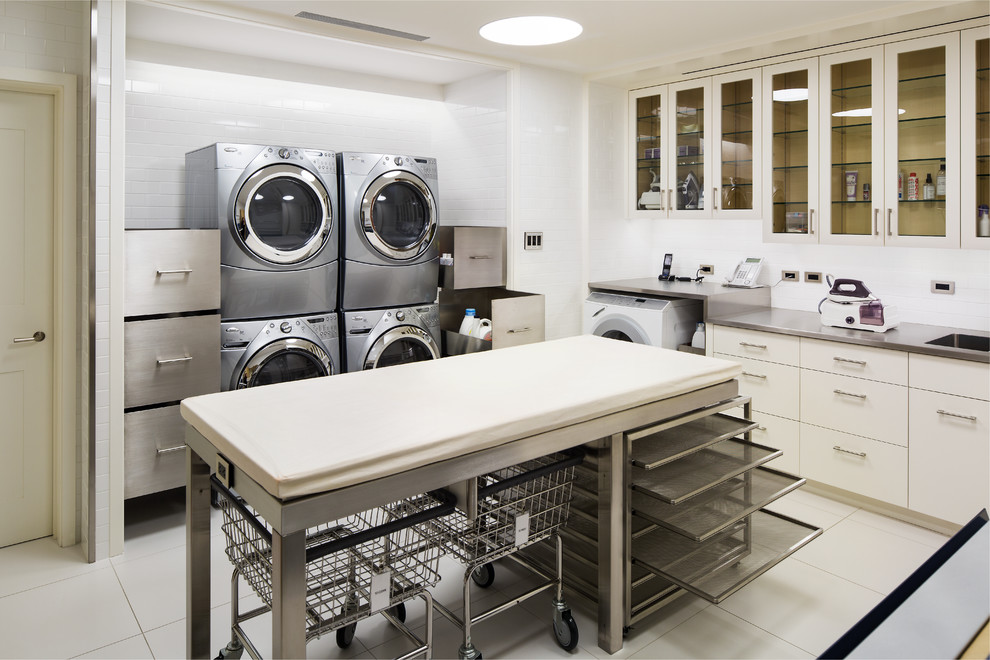 Inspiration for an expansive transitional u-shaped dedicated laundry room in New York with an undermount sink, glass-front cabinets, white cabinets, stainless steel benchtops, white walls and a stacked washer and dryer.