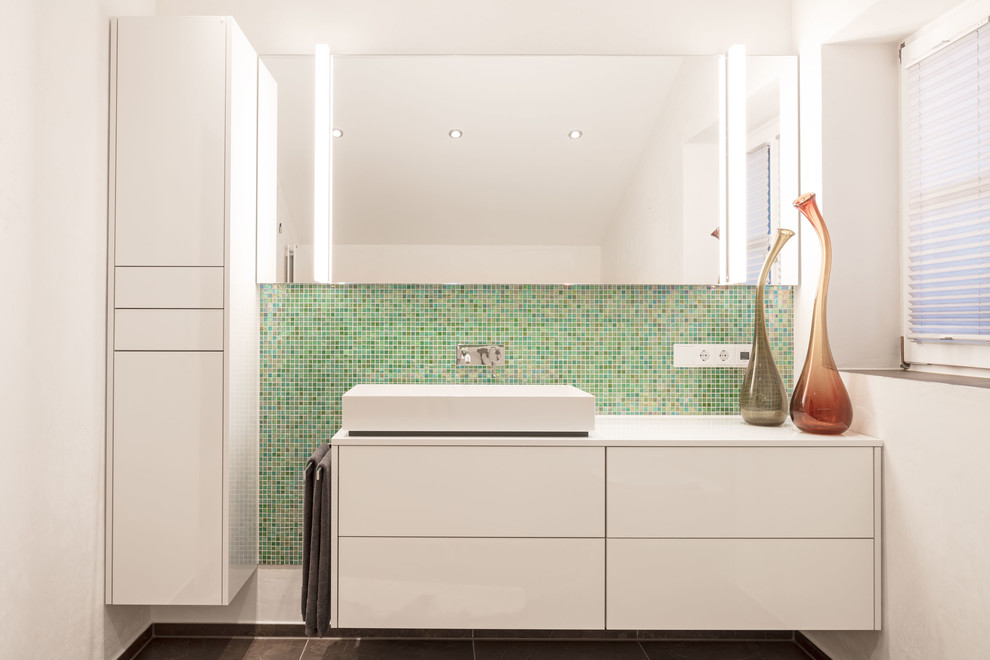 This is an example of a mid-sized contemporary bathroom in Munich with flat-panel cabinets, white cabinets, a drop-in tub, an alcove shower, a wall-mount toilet, green tile, glass tile, white walls, limestone floors, a vessel sink, stainless steel benchtops, brown floor, a hinged shower door and white benchtops.