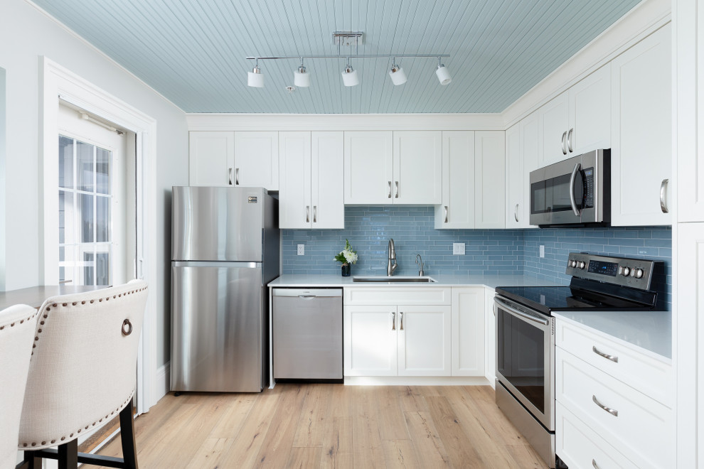Design ideas for a transitional l-shaped kitchen in Vancouver with an undermount sink, shaker cabinets, white cabinets, blue splashback, subway tile splashback, stainless steel appliances, light hardwood floors, beige floor and white benchtop.