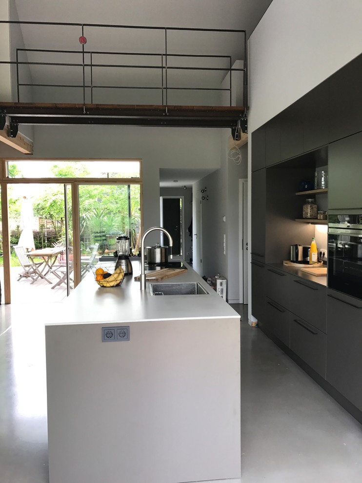 Photo of an expansive industrial galley eat-in kitchen in Other with an integrated sink, flat-panel cabinets, grey cabinets, concrete benchtops, white splashback, black appliances, concrete floors, with island, grey floor and grey benchtop.